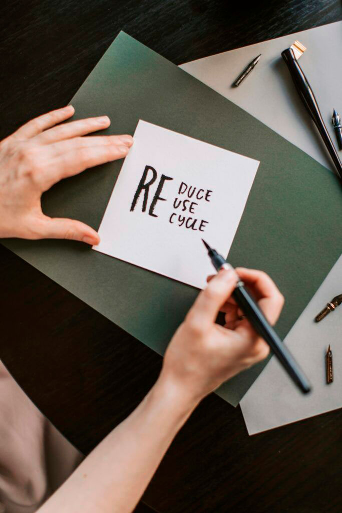 Primer plano de una mano escribiendo "Reduce, Reuse, Recycle" en una tarjeta, representando los principios de la economía circular.