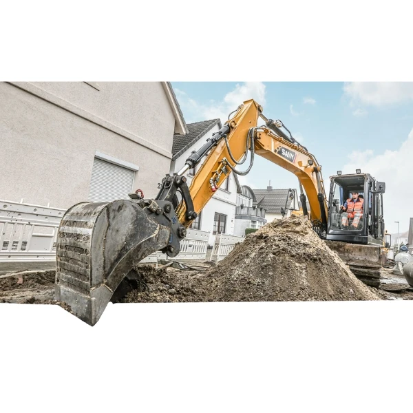 Excavadora Sany SY135C Trabajando En Obra Urbana, Cargando Tierra En Una Zanja Junto A Viviendas.